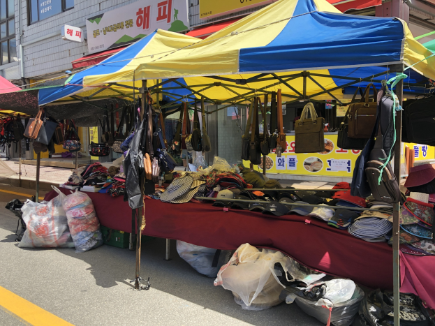 Ein Markt mit Zelten, die Taschen, Hüte und andere Artikel präsentieren, vor Gebäuden mit Fenstern und Texttafeln.