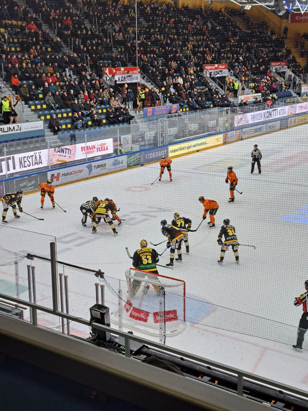 Eine Gruppe von Menschen, die Hockey auf einer Indoor-Eisfläche spielen, umgeben von einem Zaun, mit Zuschauern im Hintergrund und beleuchteter Stadionatmosphäre.