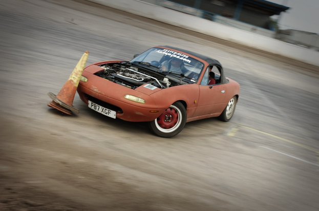 Eine Person fährt ein rotes Auto mit hoher Geschwindigkeit auf einer Rennstrecke, mit einem Verkehrskegel im Vordergrund und einem unscharfen Hintergrund.