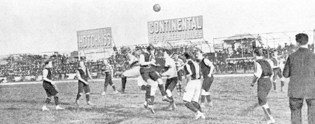 Schwarzes und weißes Bild von Männern, die auf einem Feld Fußball spielen, mit einem Ball in der Luft und Zuschauern im Hintergrund, Tafeln mit Matchdetails sichtbar.