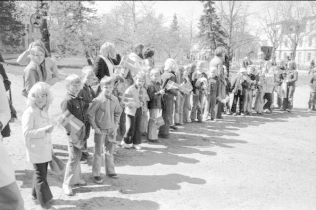 Schwarzes und weißes Bild von Menschen, die Fahnen in einer Reihe auf einer Schotterstraße während einer Protestmarsch auf dem Schulgelände halten, mit Bäumen, Gebäuden und einem klaren Himmel im Hintergrund.