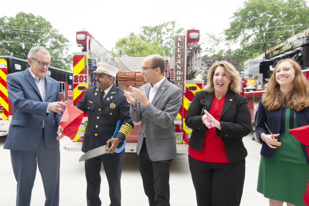 Gruppe von Menschen bei einer Eröffnungszeremonie der Alexandria Fire Department, die vor einem Feuerwehrauto klatschen und lächeln, vor einem Hintergrund mit Bäumen und einem klaren Himmel.