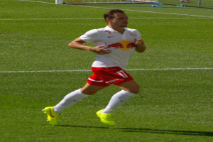 Ein Mann in einem weißen Hemd und roten Shorts rennt auf einem Fußballfeld, im Hintergrund ist ein Tor zu sehen und ein paar Menschen stehen daneben, die Fußballschuhe tragen.