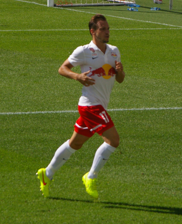 Ein Mann in einem weißen Hemd und roten Shorts rennt auf einem Fußballfeld, im Hintergrund ist ein Tor zu sehen und ein paar Menschen stehen daneben, die Fußballschuhe tragen.