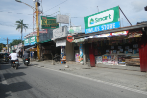 Eine belebte Straße voller Motorräder, Autos, Gebäude, Geschäfte, Schilder, Strommasten, Kabel, Straßenlaternen, Bäume und eine bewölkte Himmel.