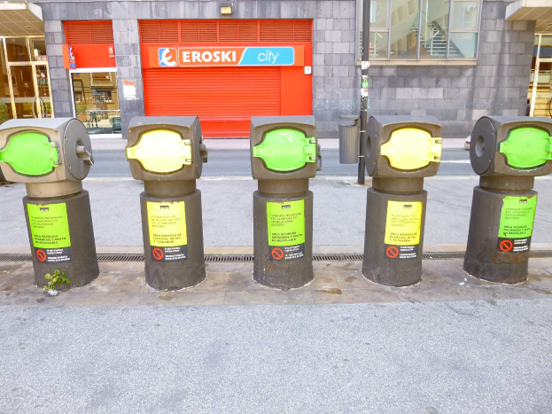 Eine Reihe von Parkautomaten auf einer Straße, mit einem Gebäude mit Glasfenstern und einem Rollladen mit Text im Hintergrund und einem nahen Müllcontainer.