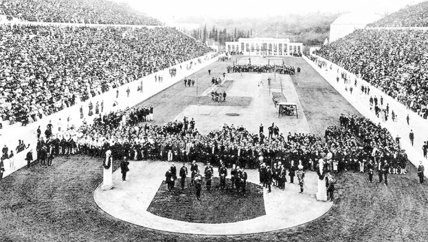 Schwarzes und weißes Foto einer großen Menschenmenge in einem Stadion während der Eröffnungszeremonie der Olympischen Spiele 1896, mit Menschen auf den Rängen sitzend und anderen auf dem Boden stehend oder sitzend, Bäume und ein Gebäude im Hintergrund sichtbar.