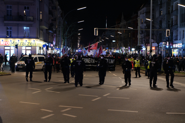 Eine Gruppe von Menschen, die über eine nächtliche Stadtstra├če gehen, mit Laternen, Geb├Ąuden, B├Ąumen, Ampeln, Fahrzeugen und einem Banner im Hintergrund.