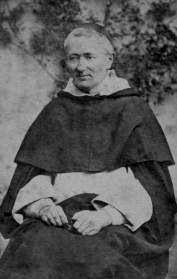 Ein altes Schwarz-Weiß-Foto eines Mannes in Priesterroben, der vor Bäumen sitzt, mit einem schwarzen Gewand, weißem Kragen und Manschetten, die Hände zum Gebet gefaltet, mit einem friedlichen Ausdruck, der von warmem Licht erhellt wird.