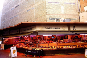 Ein belebter Markt mit verschiedenen Speisen, die unter Lichtern präsentiert werden, umgeben von Texttafeln, mit Gebäuden und einem klaren blauen Himmel im Hintergrund.