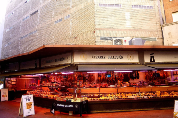 Ein belebter Markt mit verschiedenen Speisen, die unter Lichtern präsentiert werden, umgeben von Texttafeln, mit Gebäuden und einem klaren blauen Himmel im Hintergrund.
