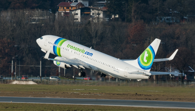 Großes Passagierflugzeug steigt von einer Flughafenrollbahn auf, umgeben von Gras, Straße, Zaun, Pfählen, Fahrzeugen, Bäumen und Gebäuden im Hintergrund.