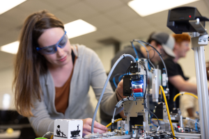 Eine Frau in Schutzbrille arbeitet an einem Roboter in einem Labor umgeben von anderen, mit Werkzeugen und Komponenten auf dem Tisch unter Deckenbeleuchtung.