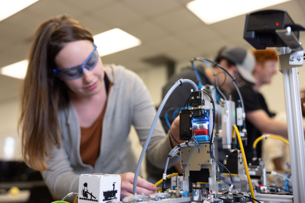 Eine Frau in Schutzbrille arbeitet an einem Roboter in einem Labor umgeben von anderen, mit Werkzeugen und Komponenten auf dem Tisch unter Deckenbeleuchtung.