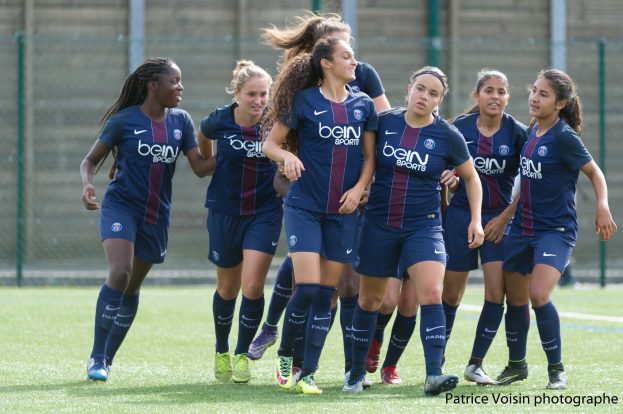 Gruppe junger Frauen, die Fußball auf einem Rasenfeld mit Maschendrahtzaun und einer Wand im Hintergrund spielen, Text in der rechten unteren Ecke lautet "Paris Saint-Germain Women's Soccer".