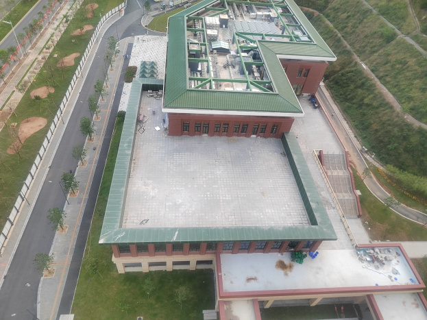 Luftaufnahme eines großen Schulgebäudes mit grüner Dachfläche, umgeben von Bäumen, Gras, Straßen, Fahrzeugen, Polen und einem Zaun.