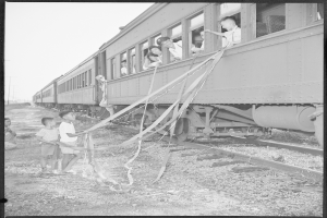 Schwarzes und weißes Foto einer Gruppe von Kindern, die neben einem Zug auf einem Bahngleis stehen, einige halten Bänder, mit Pfählen und einem klaren Himmel im Hintergrund.