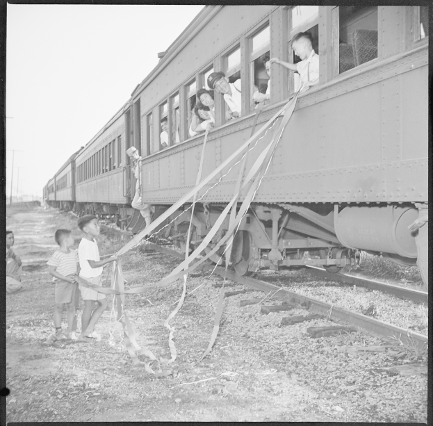 Schwarzes und weißes Foto einer Gruppe von Kindern, die neben einem Zug auf einem Bahngleis stehen, einige halten Bänder, mit Pfählen und einem klaren Himmel im Hintergrund.