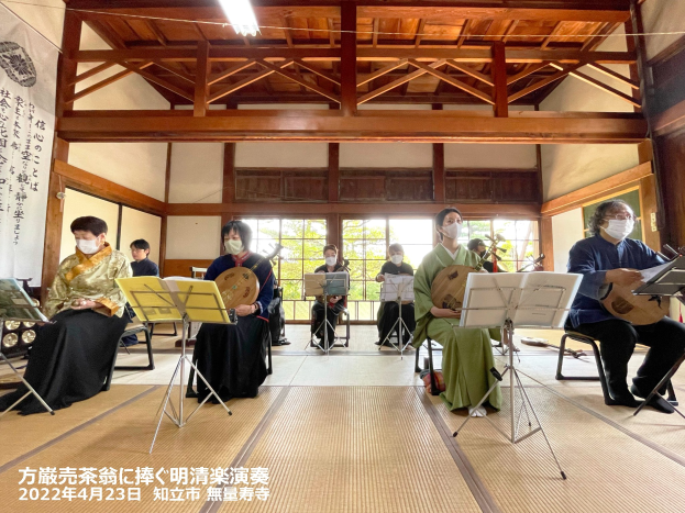Eine Gruppe von Menschen in Masken spielt Musikinstrumente in einem Raum mit Bücherständern, einem Banner, Glasfenstern, Deckenlampen und Text am unteren Rand, der die Teilnahme an einem japanischen Musikfestival anzeigt.