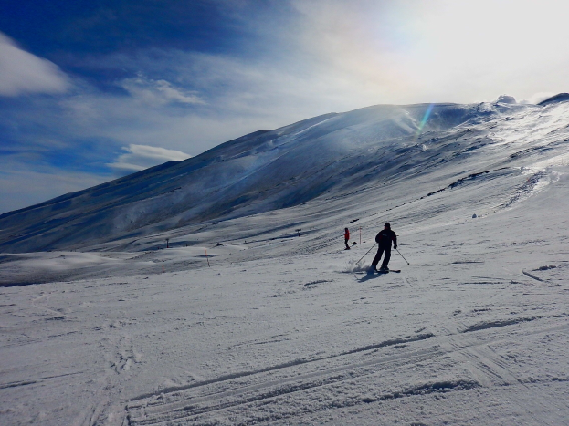 Eine Person fährt Ski eine schneebedeckte Piste hinunter mit Bergen im Hintergrund, trägt einen Helm, Handschuhe und Schuhe, hält Ski-Stöcke in der Hand, unter einem bewölkten Himmel mit glitzerndem Schnee in der Sonne.
