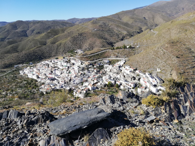 Kleines Dorf in einem Bergtal mit verstreuten Häusern, umgeben von Felsen, Pflanzen und Bäumen unter einem sichtbaren Himmel.