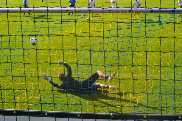 Ein Fußballtorwart, der einen Ball vor dem Tor abwehrt, vor einem Netz im Vordergrund und einigen Menschen im Hintergrund, während mehrere Fußballs auf dem Boden verstreut sind.