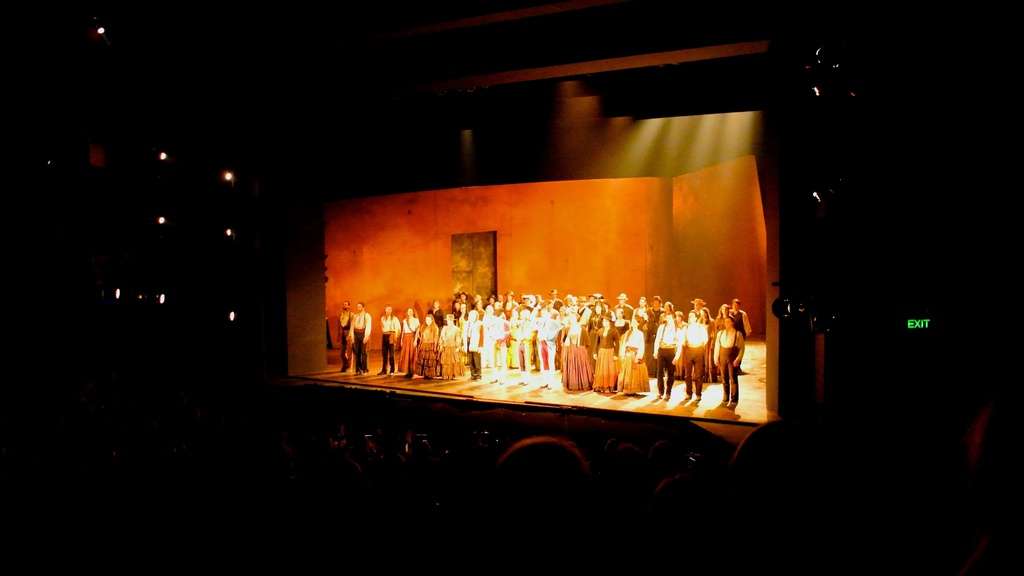 Eine Bühne mit Menschen in altertümlicher Kleidung, umgeben von Zuschauern und Scheinwerfern links.