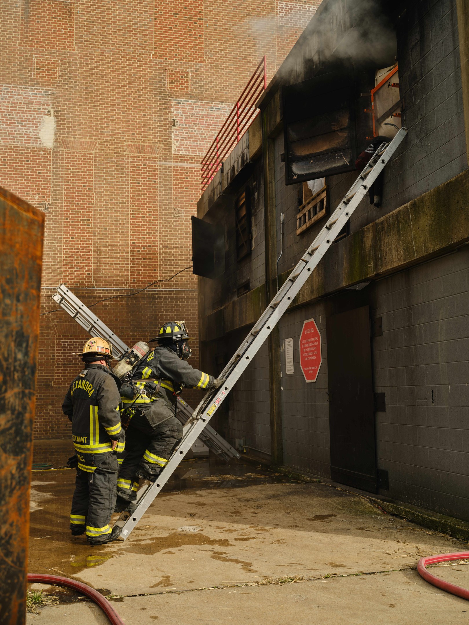 Feuerwehrleute in Helmen, die eine Leiter zu einem raucherfüllten Gebäude hinaufklettern, mit einem Metallobjekt links und Rohren am Boden.