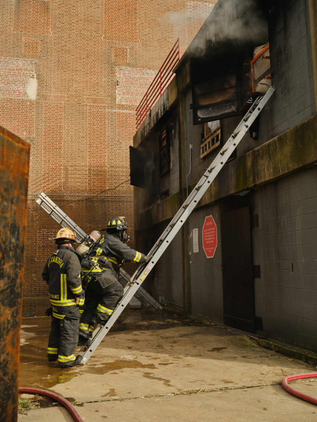 Feuerwehrleute in Helmen, die eine Leiter zu einem raucherfüllten Gebäude hinaufklettern, mit einem Metallobjekt links und Rohren am Boden.