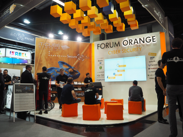 Gruppe von Menschen an einem IT-Cube Systems Forum Orange Cyber Security Stand, mit einem Anzeigebildschirm, Texttafeln, Regalen, Beschilderung und Deckenbeleuchtung.