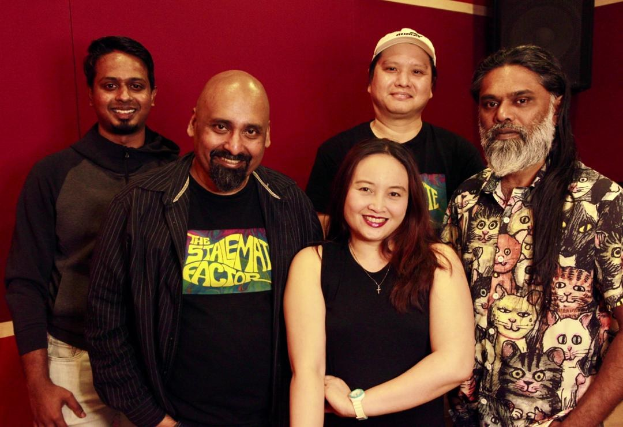 Eine Gruppe von Menschen, die vor einer roten Wand stehen und lächeln, mit einem Lautsprecher im Hintergrund, der darauf hindeutet, dass sie sich in einem Aufnahmestudio befinden und Teil einer Band zu sein scheinen.