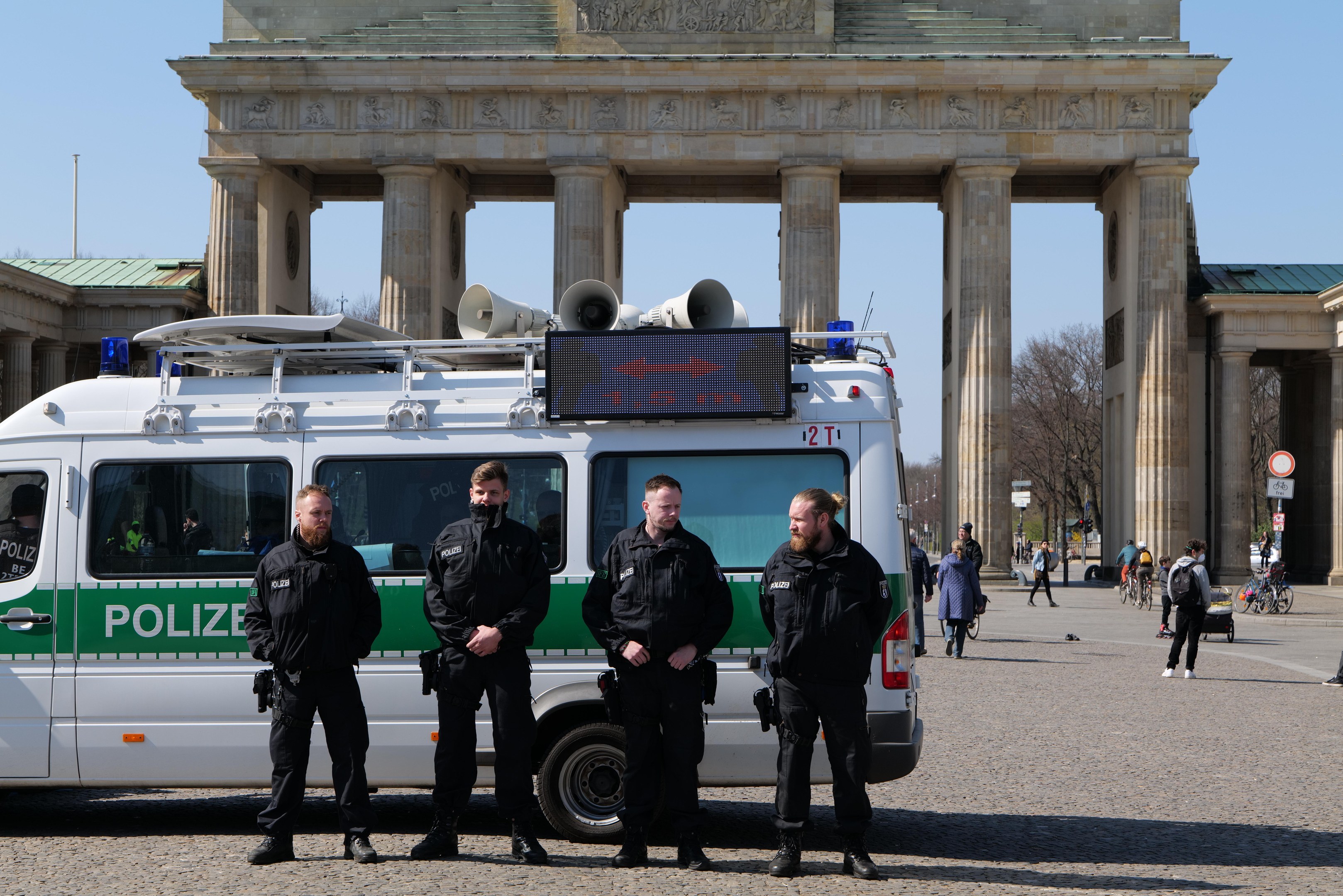 Polizeibeamte in schwarzen Uniformen stehen vor dem Brandenburger Tor in Berlin, mit einem weißen und grünen Fahrzeug im Vordergrund und Fußgängern und Radfahrern im Hintergrund.