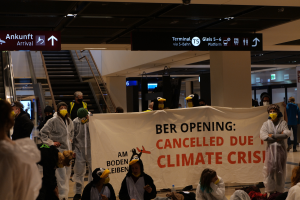 Eine Gruppe maskierter Personen mit einer Tafel, auf der "Ber Opening Due to Climate Crisis" steht, umgeben von verstreuten Taschen und Gegenständen, vor einem Hintergrund mit Rolltreppen, Texttafeln, Lichtern und Säulen.