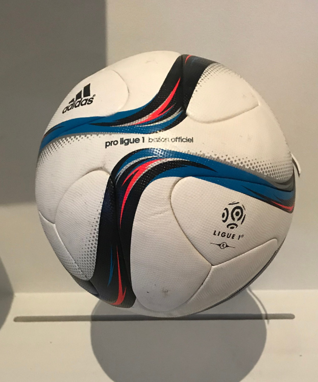 Ein weißer Fußball mit schwarzer "Ligue 1"-Aufschrift und Adidas-Logo auf einem weißen Tisch vor einem hellen weißen Hintergrund.