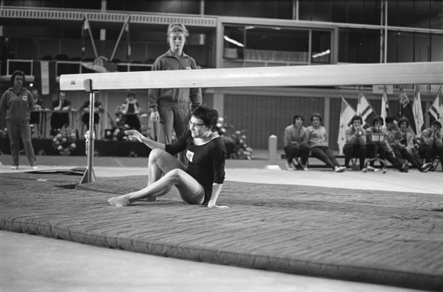 Eine Frau in einem schwarzen und weißen Turnanzug führt auf einem Schwebebalken in einer Turnhalle mit Zuschauern und Fahnen im Hintergrund auf.