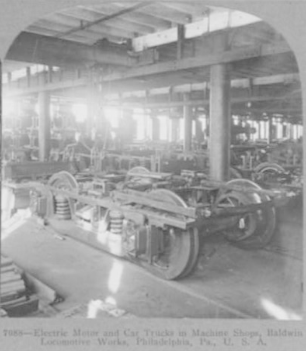 Schwar\042weißes Foto eines Elektromotors und eines Lkw in einer Fabrik mit Maschinen, S"ulen und Dach, mit Text unten.