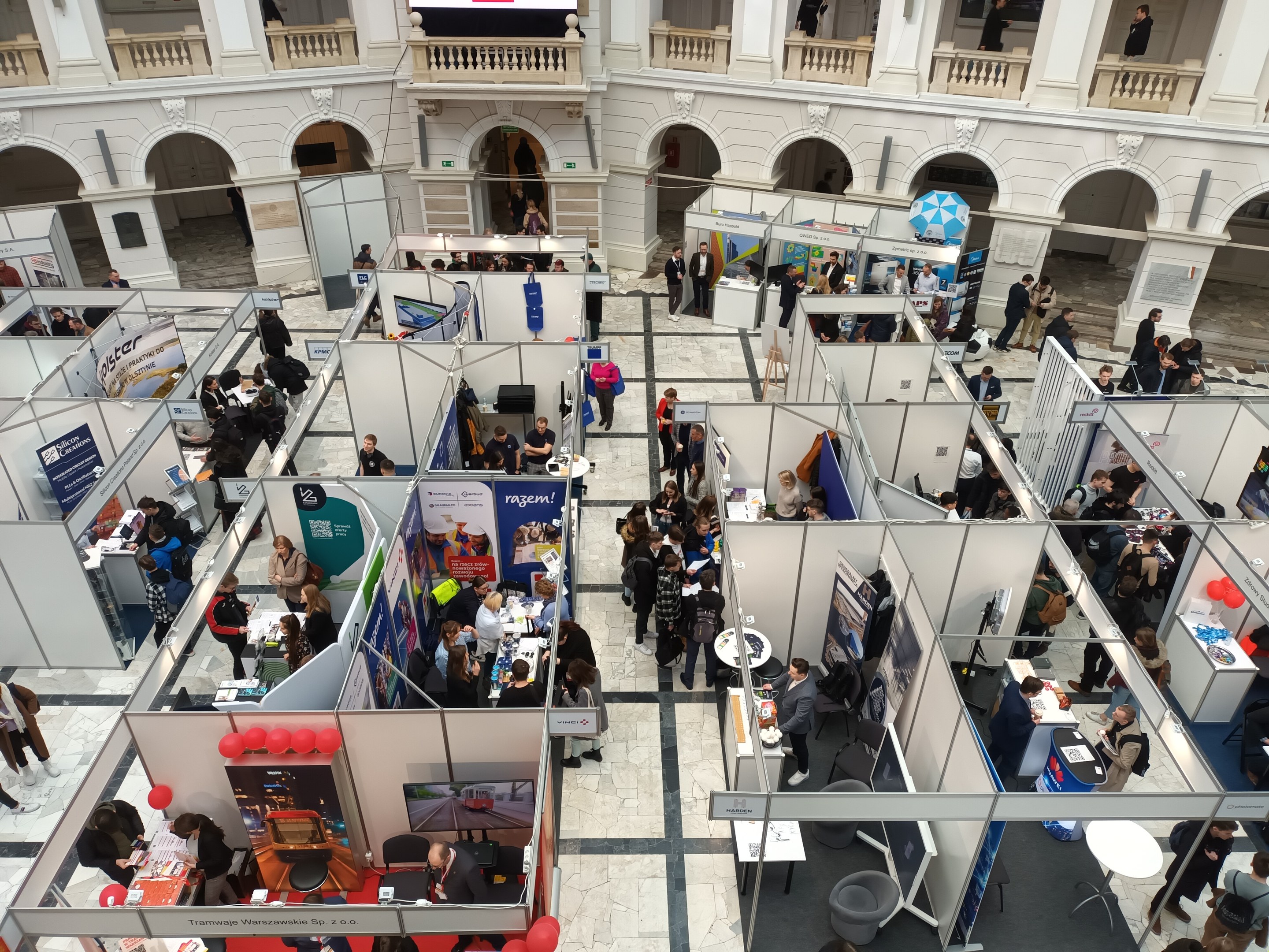 Große Menschenmenge auf einer internationalen Messe mit Ständen, Bannern, Tischen, Stühlen, Luftballons und einem Gebäude mit Bögen, Säulen, Geländern und einem Bildschirm im Hintergrund.