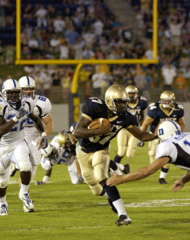 Fußballspieler in Bewegung mit dem Ball, verfolgt von Gegnern in Sportbekleidung und Helmen, mit unscharfen Zuschauern und Stadionelementen im Hintergrund und Text unten.