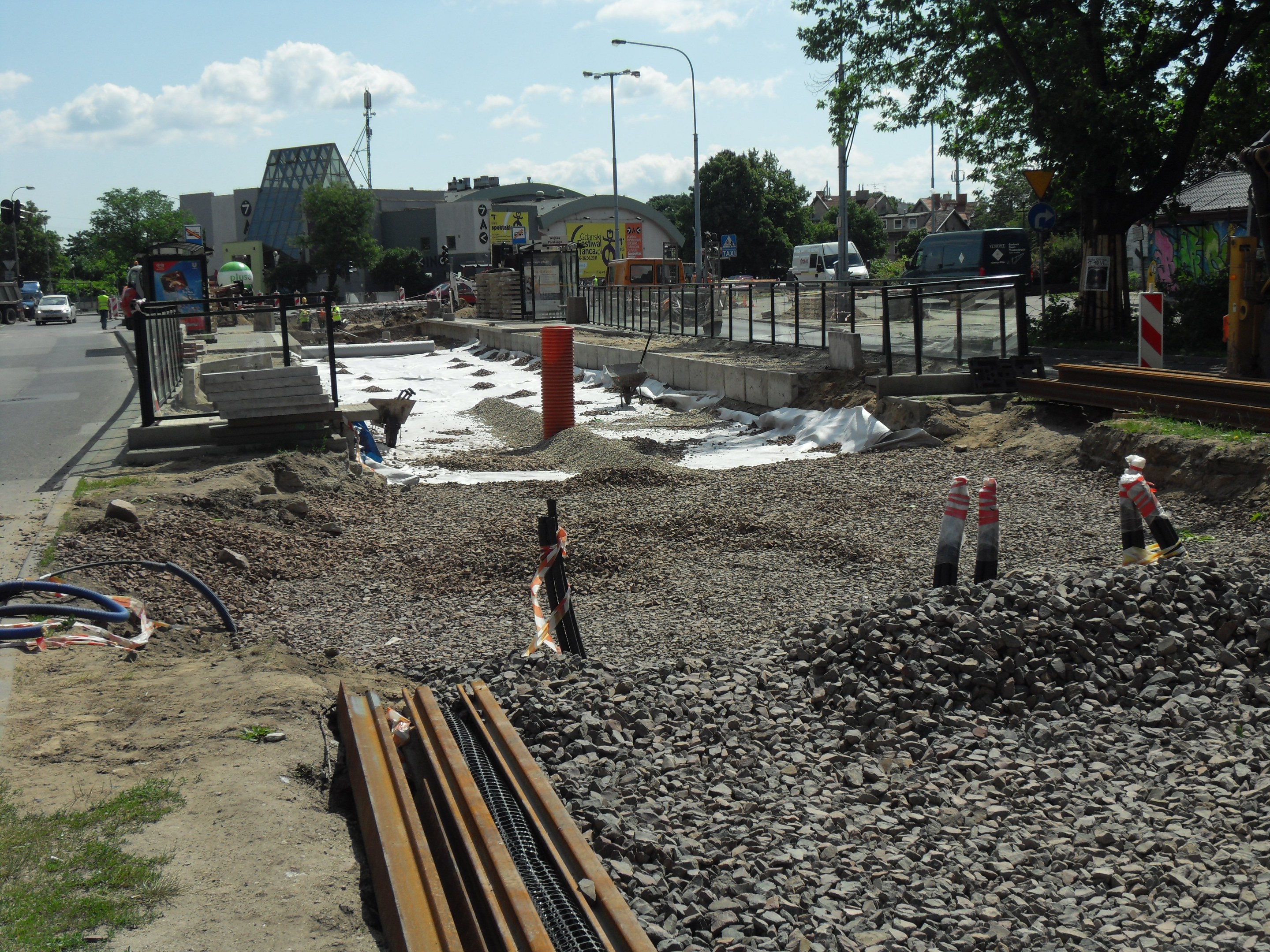 Baustelle mit verschiedenen Materialien und Geräten am Straßenrand, Fahrzeuge und Gebäude im Hintergrund unter einem bewölkten Himmel.