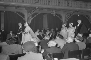 Schwarzes und weißes Foto einer Hochzeitszeremonie in einem gro├čen Raum mit sitzenden und stehenden G├Ąsten, einige halten Blumenstr├Ąu├če, Tische mit Tassen und Gegenst├Ąnden und Vorh├Ąnge mit Deckenleuchten im Hintergrund.