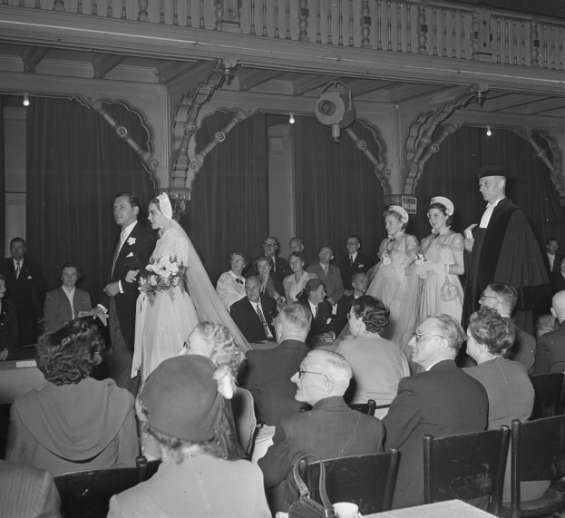 Schwarzes und weißes Foto einer Hochzeitszeremonie in einem gro├čen Raum mit sitzenden und stehenden G├Ąsten, einige halten Blumenstr├Ąu├če, Tische mit Tassen und Gegenst├Ąnden und Vorh├Ąnge mit Deckenleuchten im Hintergrund.