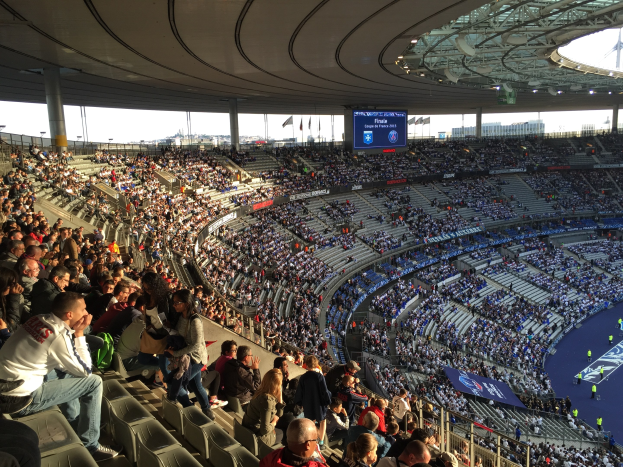 Große Menge im Stadion bei einem Fußballspiel, mit einer Bühne rechts, Fahnen, Stangen, einem Bildschirm und der Allianz Arena in München, Deutschland im Hintergrund.