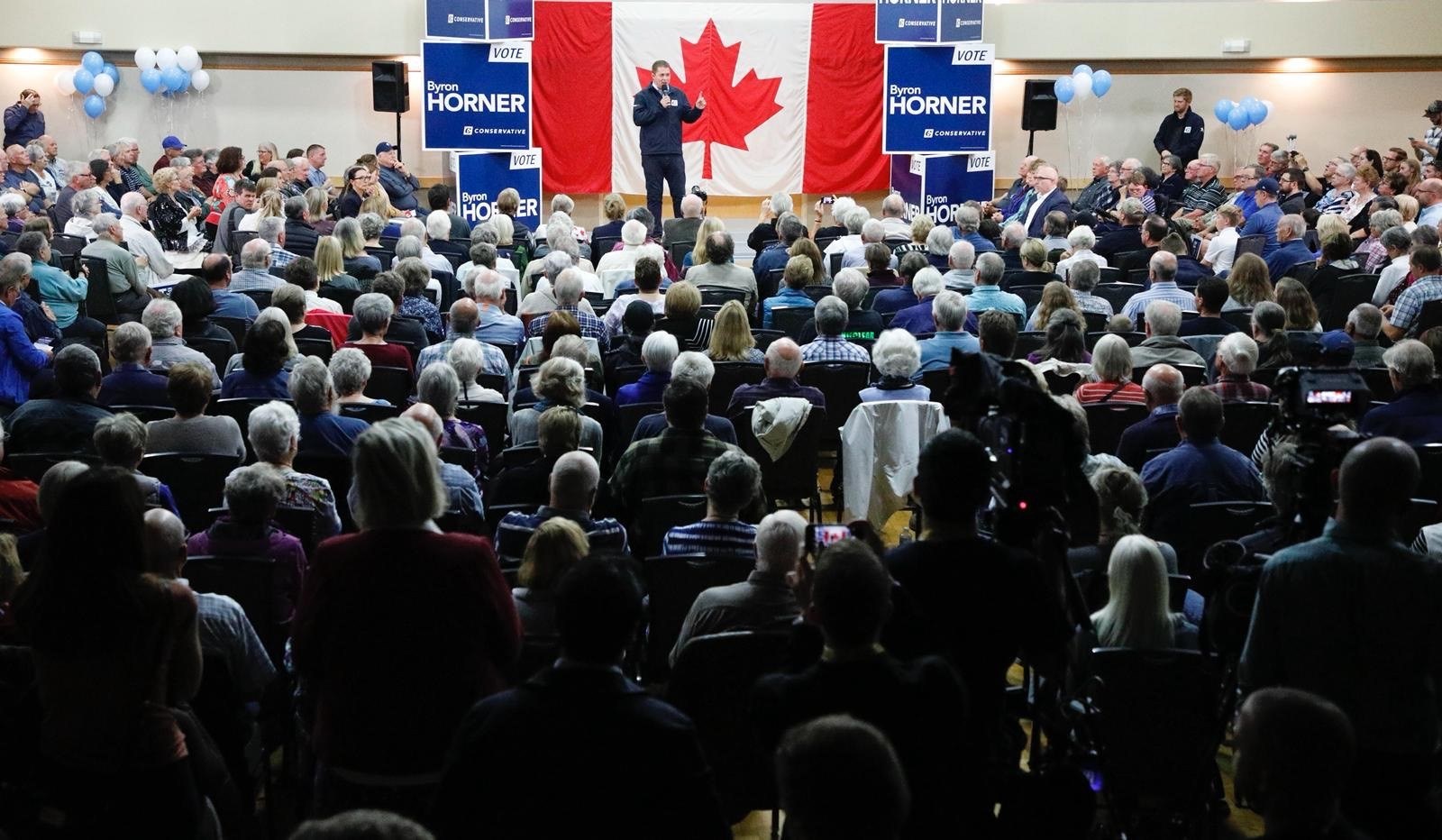 Eine große Menschenmenge sitzt auf Stühlen vor einer kanadischen Flagge, ein Mann hält ein Mikrofon auf einem Podium, umgeben von Bannern, Luftballons und Lautsprechern auf dem Byron Horner Conference.