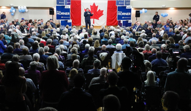 Eine große Menschenmenge sitzt auf Stühlen vor einer kanadischen Flagge, ein Mann hält ein Mikrofon auf einem Podium, umgeben von Bannern, Luftballons und Lautsprechern auf dem Byron Horner Conference.