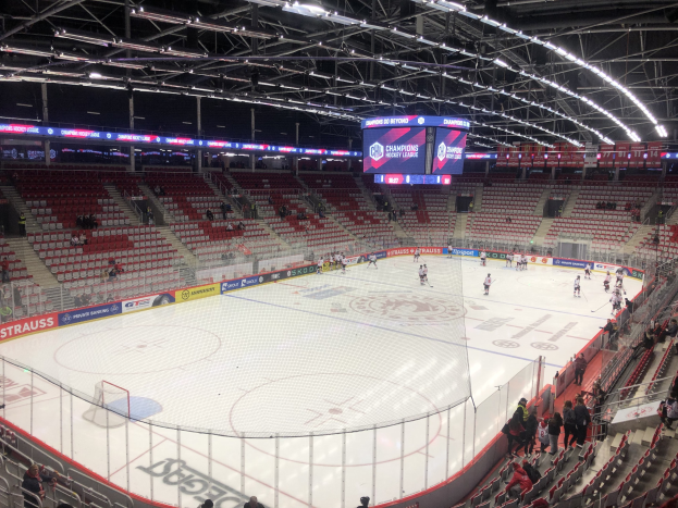 Hockey-Spiel in einer großen Arena mit Zuschauern, eine Eisfläche umgeben von einem Zaun, ein Display mit "St. Louis Blues gegen Detroit Red Wings", Metallrahmen, Deckenleuchten und ein Dach.