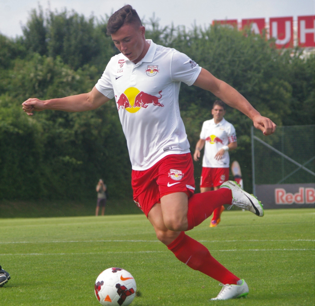 Ein professioneller Fußballer tritt einen Ball auf einem grünen Feld mit Bäumen und einem klaren blauen Himmel im Hintergrund, in der Nähe einer Tafel mit der Aufschrift "RB Leipzig."