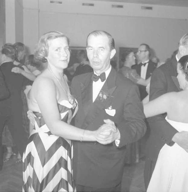 Zwei Personen, ein Mann in einem schwarzen Blazer und weißem Hemd mit schwarzer Hose und eine Frau in einem unbekannten Outfit, tanzen zusammen auf einer formellen Veranstaltung, halten sich an den Händen und wiegen sich im Takt gegen eine Wand im Hintergrund.