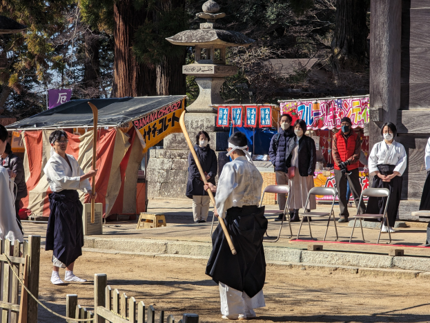 Eine Gruppe von Menschen in formeller Kleidung, einige tragen Masken und halten Holzstöcke, versammeln sich draußen vor einem traditionellen Gebäude während einer Zeremonie in Kyoto, mit Stühlen, Bannern, Bäumen und einem klaren Himmel.