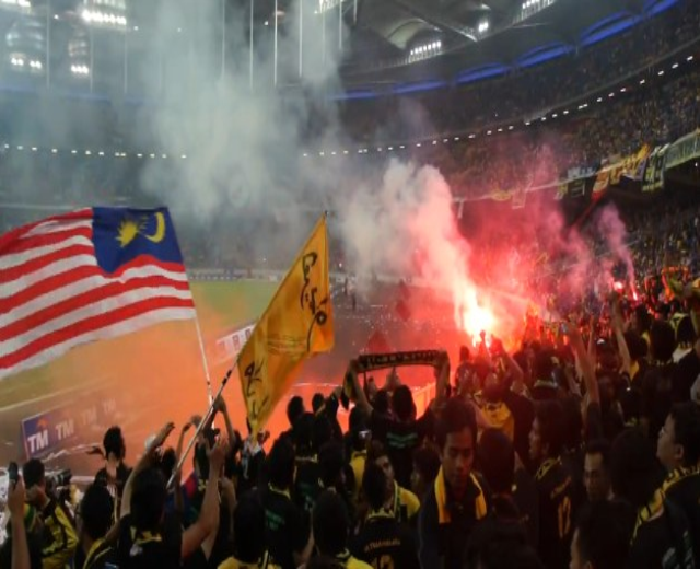 Eine große Menge Menschen in einem Stadion mit Feuerwerk in der Luft, einige halten Fahnen und eine Person hält eine Kamera, mit Rauch im Hintergrund und Schildern mit der Aufschrift "Malaysia vs Malaysia Live-Streaming."