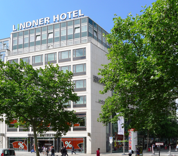 Großes modernes Hotelgebäude mit Glasfenstern, der Name "Lindner" oben drauf, umgeben von Bäumen, Fahrzeugen und Fußgängern unter einem sichtbaren Himmel.